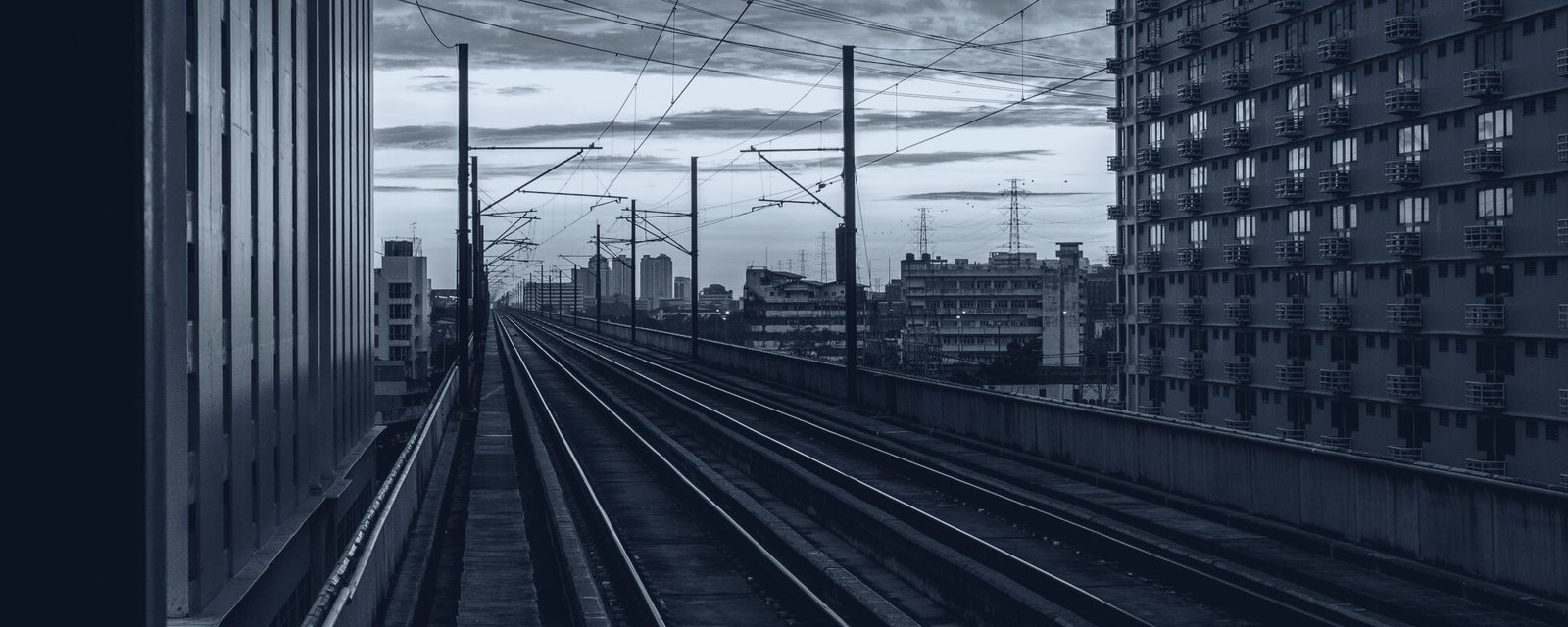 Elevated twin-track rail corridor with overhead line masts receding to a city skyline, slate duotone — atmospheric imagery